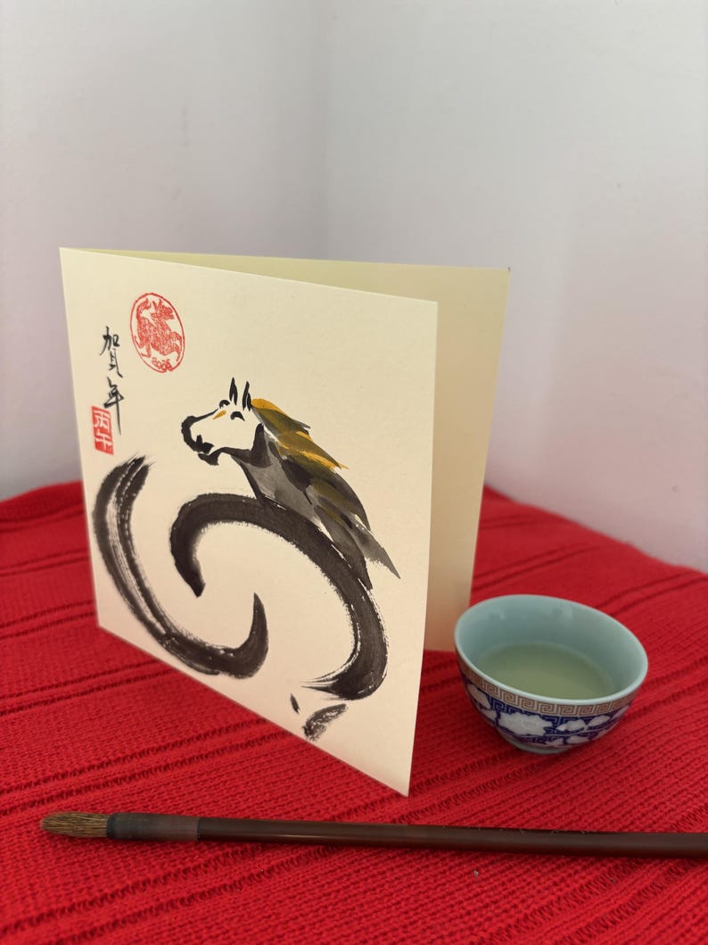 Pode incluir: Um cart&atilde;o de felicita&ccedil;&otilde;es creme com uma ilustra&ccedil;&atilde;o de cavalo preto e cinza e caracteres japoneses. Uma pequena ch&aacute;vena de ch&aacute; azul e branca e um pincel numa superf&iacute;cie vermelha.