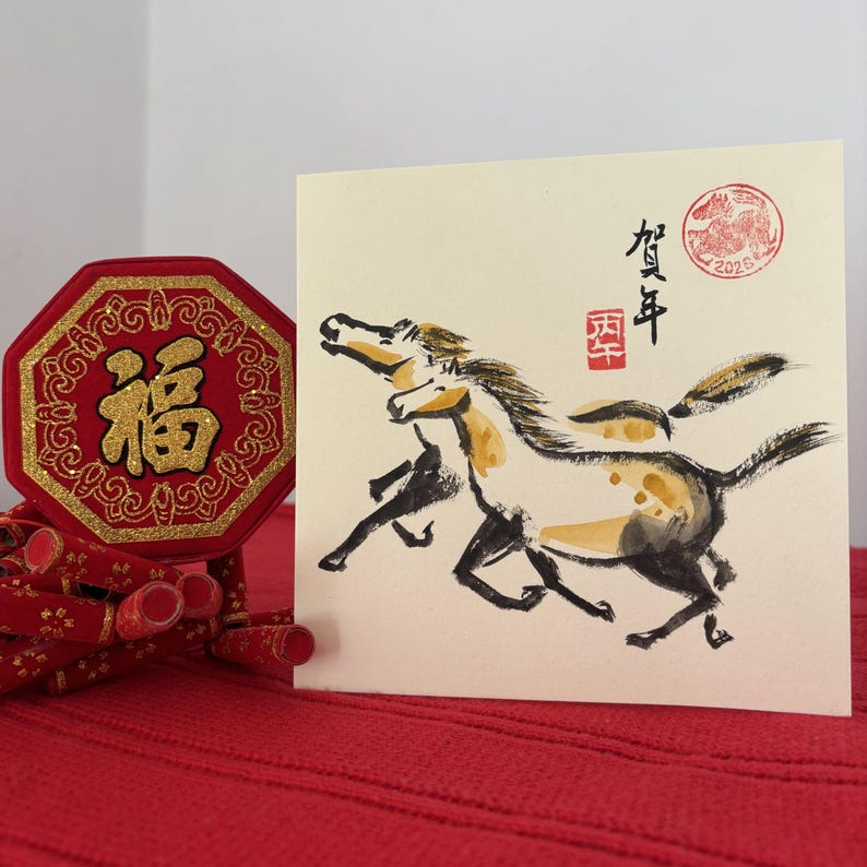 May include: A square greeting card with a watercolor painting of two running horses in black, white, and gold. The card has Chinese characters and a red stamp. A red, gold, and black decorative item is on the left.