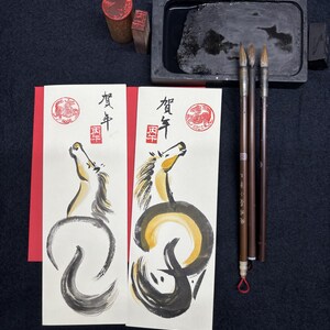 K&ouml;nnte beinhalten: Japanisches Kalligraphie-Set mit zwei illustrierten Karten mit Pferde-Design, Stempeln, Tinte, Pinseln und einem Tintenstein. Die Karten haben japanischen Text und ein rotes Siegel. Die Pferdeillustrationen sind in schwarzer und goldener Tinte.