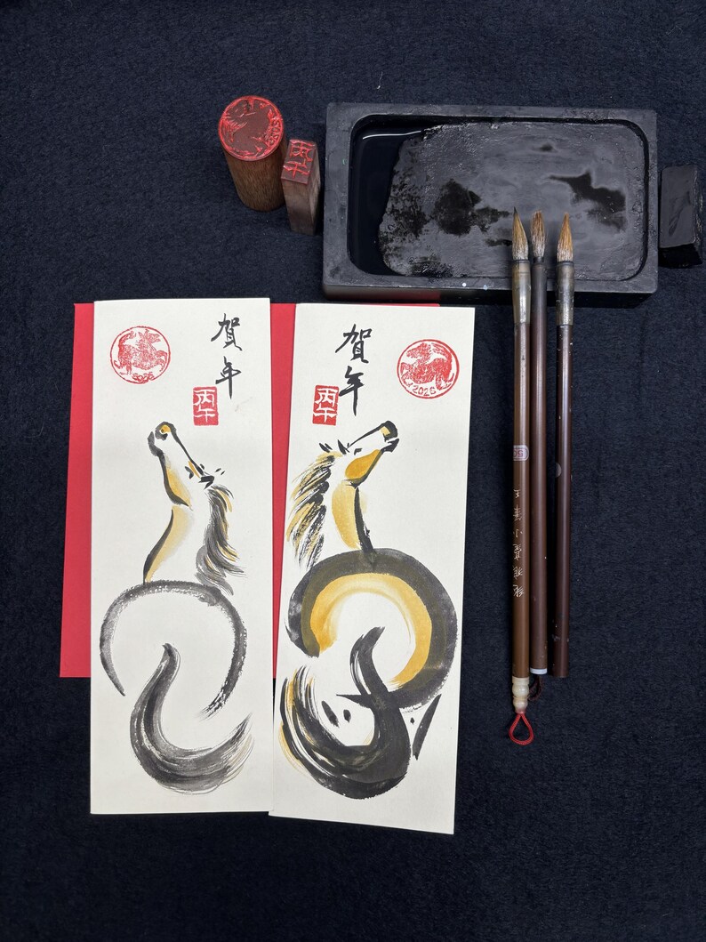 K&ouml;nnte beinhalten: Japanisches Kalligraphie-Set mit zwei illustrierten Karten mit Pferde-Design, Stempeln, Tinte, Pinseln und einem Tintenstein. Die Karten haben japanischen Text und ein rotes Siegel. Die Pferdeillustrationen sind in schwarzer und goldener Tinte.