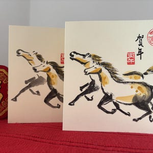 May include: Two greeting cards with watercolor paintings of running horses. The horses are depicted in black, white, and yellow, with a red stamp and Chinese characters. A red, gold-trimmed decorative item is on the left.