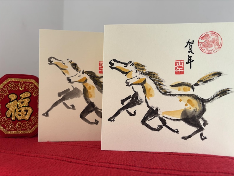 May include: Two greeting cards with watercolor paintings of running horses. The horses are depicted in black, white, and yellow, with a red stamp and Chinese characters. A red, gold-trimmed decorative item is on the left.