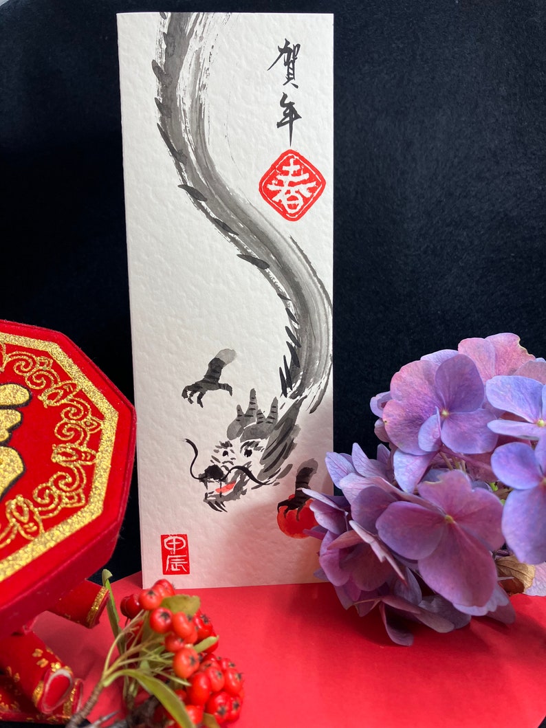 Puede incluir: Una pintura de tinta negra de un drag&oacute;n en una tarjeta blanca. El drag&oacute;n est&aacute; enroscado y tiene un c&iacute;rculo rojo con caracteres chinos en &eacute;l. La tarjeta est&aacute; rodeada de flores rojas y moradas.