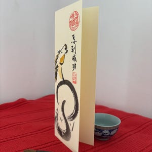 K&ouml;nnte beinhalten: Eine gefaltete cremefarbene Gru&szlig;karte mit einer Pferdeillustration in schwarzer Tinte und gelben Akzenten. Die Karte zeigt chinesische Kalligraphie und einen roten runden Stempel. Eine kleine blau-wei&szlig;e Schale und ein Kalligraphiepinsel liegen auf einer roten Oberfl&auml;che.