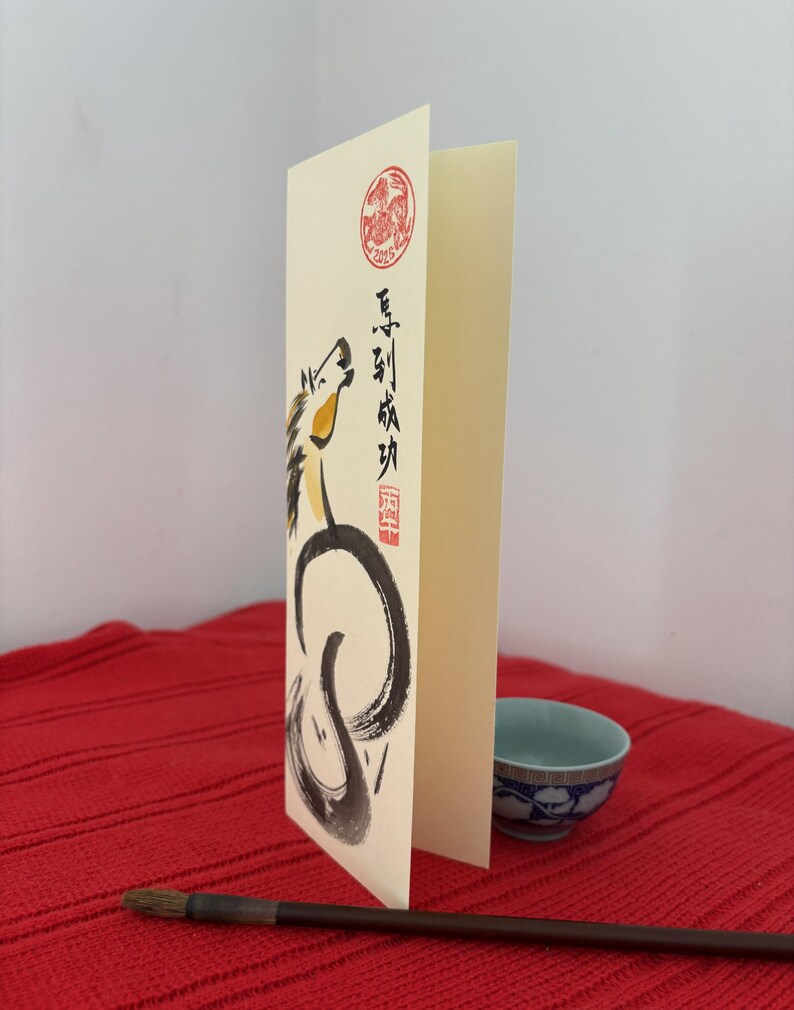 K&ouml;nnte beinhalten: Eine gefaltete cremefarbene Gru&szlig;karte mit einer Pferdeillustration in schwarzer Tinte und gelben Akzenten. Die Karte zeigt chinesische Kalligraphie und einen roten runden Stempel. Eine kleine blau-wei&szlig;e Schale und ein Kalligraphiepinsel liegen auf einer roten Oberfl&auml;che.