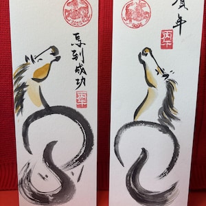 K&ouml;nnte beinhalten: Zwei rechteckige Karten mit japanischer Kalligraphie und Pferdeillustrationen. Jede Karte zeigt ein stilisiertes Pferd mit schwarzer M&auml;hne und Schweif sowie einem gelben und braunen Kopf. Rote Stempel und japanischer Text sind ebenfalls vorhanden.