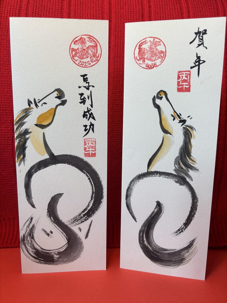 K&ouml;nnte beinhalten: Zwei rechteckige Karten mit japanischer Kalligraphie und Pferdeillustrationen. Jede Karte zeigt ein stilisiertes Pferd mit schwarzer M&auml;hne und Schweif sowie einem gelben und braunen Kopf. Rote Stempel und japanischer Text sind ebenfalls vorhanden.