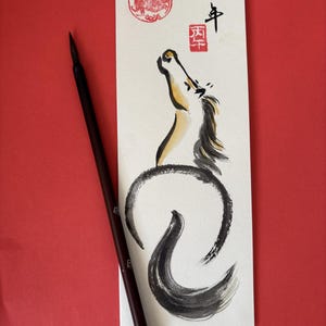K&ouml;nnte beinhalten: Ein japanisches Kalligraphie-Kunstwerk mit einer Pferdeillustration in schwarzer und goldener Tinte. Das Kunstwerk enth&auml;lt japanische Schriftzeichen und einen roten Stempel. Ein dunkelbrauner Kalligraphiepinsel liegt auf rotem Hintergrund.