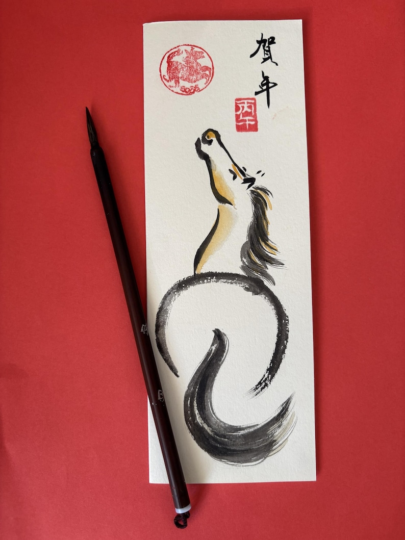 K&ouml;nnte beinhalten: Ein japanisches Kalligraphie-Kunstwerk mit einer Pferdeillustration in schwarzer und goldener Tinte. Das Kunstwerk enth&auml;lt japanische Schriftzeichen und einen roten Stempel. Ein dunkelbrauner Kalligraphiepinsel liegt auf rotem Hintergrund.