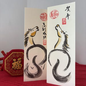 K&ouml;nnte beinhalten: Zwei cremefarbene Gru&szlig;karten mit Tuscheillustrationen von Pferden. Die Pferde sind schwarz und goldfarben, mit Kalligraphie und roten Stempeln. Ein rotes und goldenes Dekorationsobjekt befindet sich im Vordergrund.