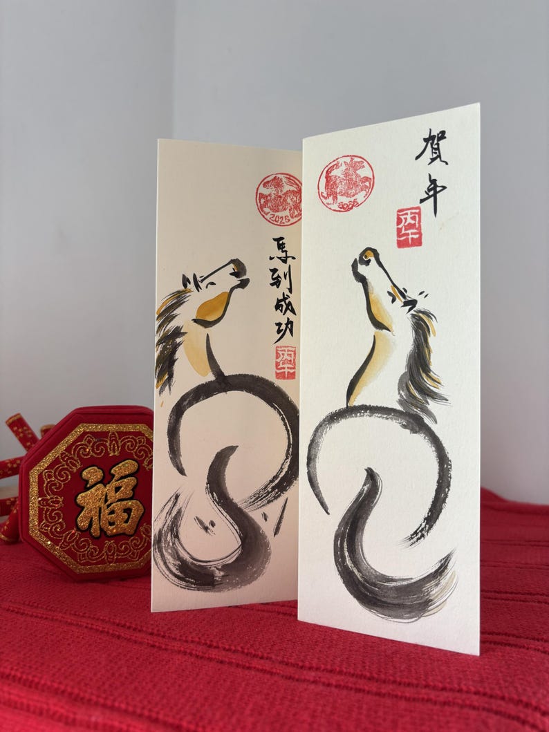 K&ouml;nnte beinhalten: Zwei cremefarbene Gru&szlig;karten mit Tuscheillustrationen von Pferden. Die Pferde sind schwarz und goldfarben, mit Kalligraphie und roten Stempeln. Ein rotes und goldenes Dekorationsobjekt befindet sich im Vordergrund.