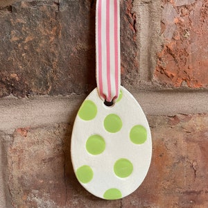 May include: White ceramic Easter egg ornament with green polka dots and a pink and white striped ribbon for hanging.