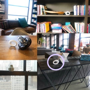 May include: A small, smooth, white oval-shaped decorative object with a hollow center. The object is shown in various settings, including on a wooden table, on a bookshelf, on a window sill, and on a dark console table next to a white geometric star.