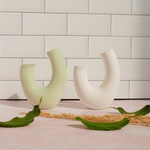 Minimalist product photography props showcasing a set of modern U-shaped plaster arch vases. The handmade concrete styling blocks feature an elegant, asymmetrical curved ring silhouette. Included are a soft, pale sage green arch and a clean white plaster riser. Standing on a pink marble countertop in front of a white subway tile backsplash, they are styled with fresh green leaves and dried grass props under bright, direct sunlight that casts dramatic shadows to the left.