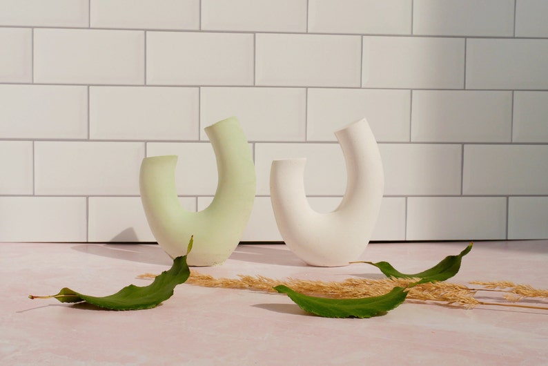 Minimalist product photography props showcasing a set of modern U-shaped plaster arch vases. The handmade concrete styling blocks feature an elegant, asymmetrical curved ring silhouette. Included are a soft, pale sage green arch and a clean white plaster riser. Standing on a pink marble countertop in front of a white subway tile backsplash, they are styled with fresh green leaves and dried grass props under bright, direct sunlight that casts dramatic shadows to the left.