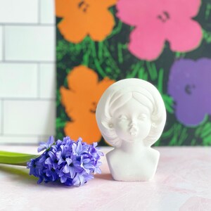 May include: White plaster bust of a woman with a closed mouth and eyes, wearing a headband. The bust is sitting on a pink surface in front of a colorful abstract painting.
