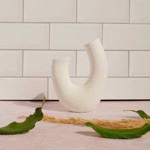 Minimalist product photography prop showcasing a single modern U-shaped plaster arch vase. The handmade white concrete styling block features an elegant, asymmetrical curved ring silhouette. Standing upright on a pink marble countertop in front of a white subway tile backsplash, the textured riser is styled with loose green leaves and dried grass under bright, direct sunlight that casts a dramatic shadow to its left. A sleek, aesthetic display stand ideal for jewelry staging.