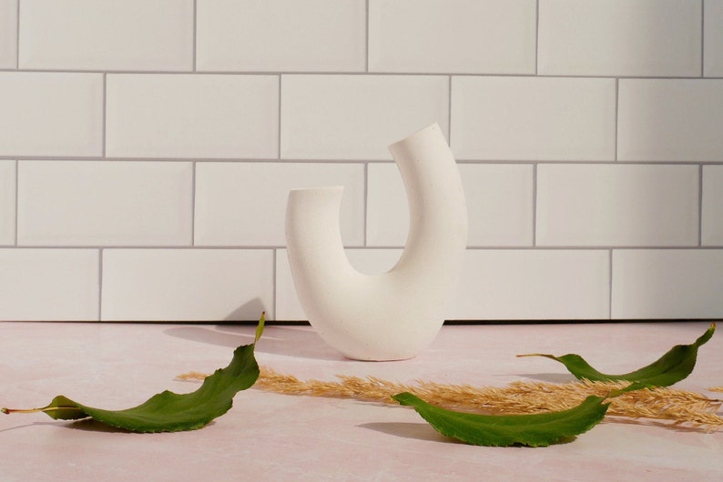 Minimalist product photography prop showcasing a single modern U-shaped plaster arch vase. The handmade white concrete styling block features an elegant, asymmetrical curved ring silhouette. Standing upright on a pink marble countertop in front of a white subway tile backsplash, the textured riser is styled with loose green leaves and dried grass under bright, direct sunlight that casts a dramatic shadow to its left. A sleek, aesthetic display stand ideal for jewelry staging.