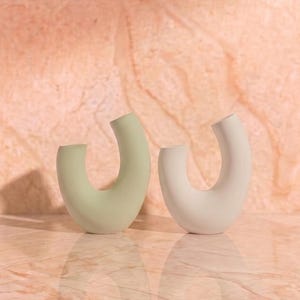 May include: Two U-shaped vases standing side by side. One vase is a soft sage green color, and the other is a matte off-white. Both vases feature a smooth, curved silhouette with an open center, creating a distinct arch shape that is uniform in design.