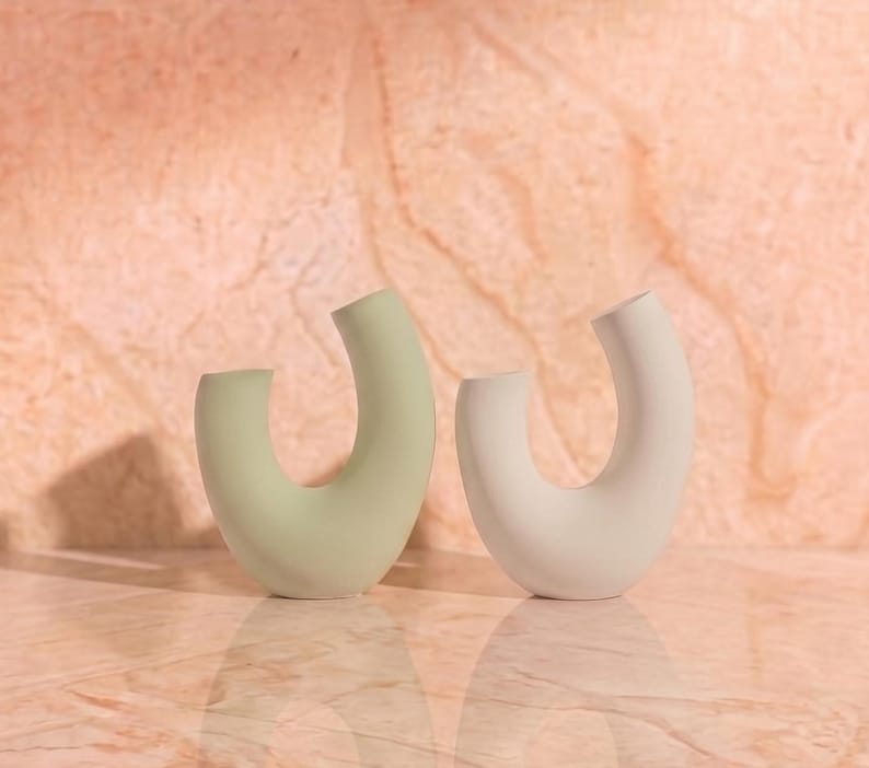 Minimalist product photography props showcasing a set of modern U-shaped plaster arch vases. The handmade concrete styling blocks feature an elegant, asymmetrical curved ring silhouette. Included are a soft pale sage green arch and a clean white plaster riser. Standing on a polished peach marble surface against a warm, textured stone background, they catch soft natural sunlight that casts subtle shadows. A sleek display stand set ideal for jewelry, cosmetics, or home fragrance staging.