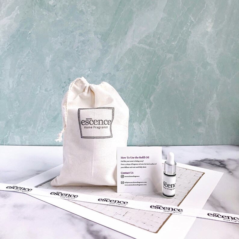May include: A beige drawstring cotton pouch with the black text "escence Home Fragrance" printed on the front, accompanied by a small white instruction card and a clear glass dropper bottle with a white cap and "escence" label.