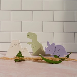 May include: A set of three wooden toy figurines, including a green dinosaur, a purple triceratops, and two white mountains. The figurines are on a white tiled background.