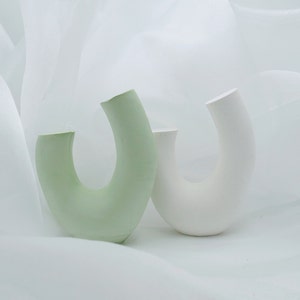 Minimalist product photography props showcasing a set of modern U-shaped plaster arch vases. The handmade concrete styling blocks feature an elegant, asymmetrical curved ring silhouette. Included are a soft, pale sage green arch and a clean white plaster riser. They are nestled inside a soft, flowing white sheer chiffon fabric backdrop with elegant waves, creating an airy, dreamlike aesthetic. A contemporary display stand set perfect for jewelry staging or home fragrance styling.