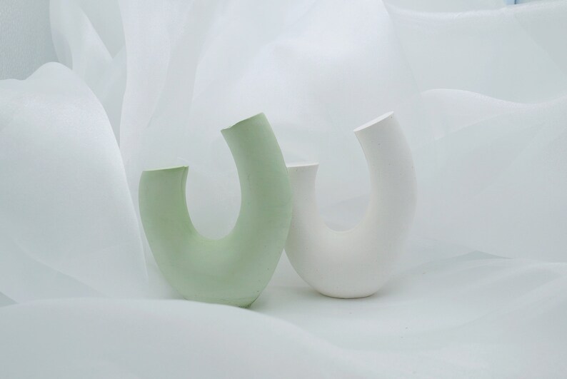 Minimalist product photography props showcasing a set of modern U-shaped plaster arch vases. The handmade concrete styling blocks feature an elegant, asymmetrical curved ring silhouette. Included are a soft, pale sage green arch and a clean white plaster riser. They are nestled inside a soft, flowing white sheer chiffon fabric backdrop with elegant waves, creating an airy, dreamlike aesthetic. A contemporary display stand set perfect for jewelry staging or home fragrance styling.