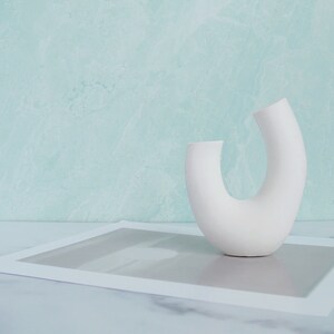 Minimalist product photography prop showcasing a single modern U-shaped plaster arch vase. The handmade white concrete styling block features an elegant, asymmetrical curved ring silhouette. It stands on a white marble surface over a gray abstract art print card, set against a soft, textured pastel teal wall background. A sleek, contemporary display riser ideal for showcasing aesthetic items, jewelry, or home fragrance styling.