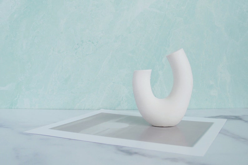 Minimalist product photography prop showcasing a single modern U-shaped plaster arch vase. The handmade white concrete styling block features an elegant, asymmetrical curved ring silhouette. It stands on a white marble surface over a gray abstract art print card, set against a soft, textured pastel teal wall background. A sleek, contemporary display riser ideal for showcasing aesthetic items, jewelry, or home fragrance styling.