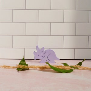 May include: A purple wooden dinosaur toy in the shape of a triceratops. The toy is sitting on a white tiled surface with a few blades of dried grass and green leaves.