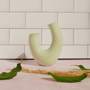 Minimalist product photography prop showcasing a single modern U-shaped plaster arch vase. The handmade concrete styling block features an elegant, asymmetrical curved ring silhouette finished in a soft, pale sage green tone. Standing upright on a pink marble countertop in front of a white subway tile backsplash, the textured riser is styled with loose green leaves and dried grass under bright, direct sunlight. A sleek, aesthetic display stand ideal for jewelry staging.