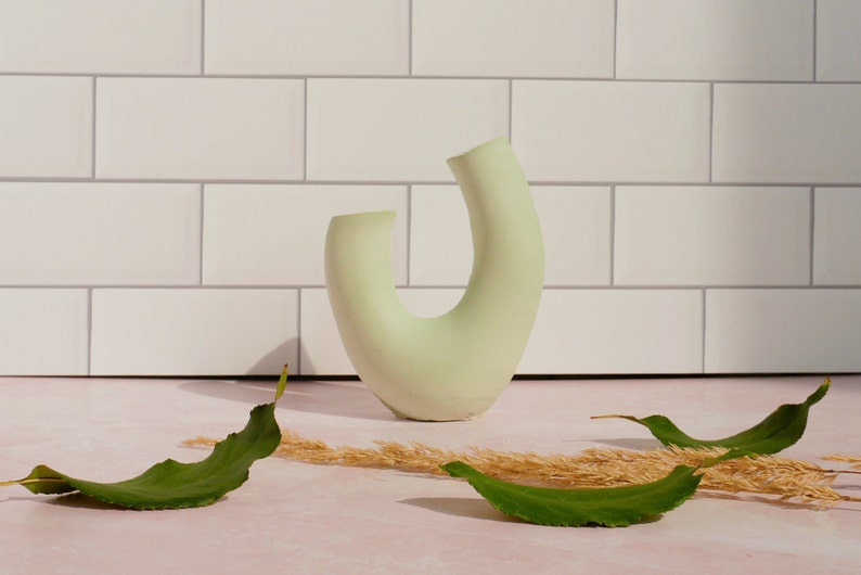 Minimalist product photography prop showcasing a single modern U-shaped plaster arch vase. The handmade concrete styling block features an elegant, asymmetrical curved ring silhouette finished in a soft, pale sage green tone. Standing upright on a pink marble countertop in front of a white subway tile backsplash, the textured riser is styled with loose green leaves and dried grass under bright, direct sunlight. A sleek, aesthetic display stand ideal for jewelry staging.