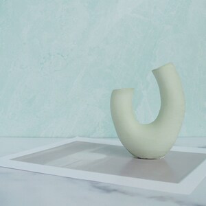 Minimalist product photography prop showcasing a single modern U-shaped plaster arch vase. The handmade concrete styling block features an elegant, asymmetrical curved ring silhouette in a soft, pale sage green. It stands on a white marble surface over a gray abstract art print card, set against a soft, textured pastel teal wall background. A sleek, contemporary display riser ideal for showcasing aesthetic items, jewelry, or home fragrance styling.