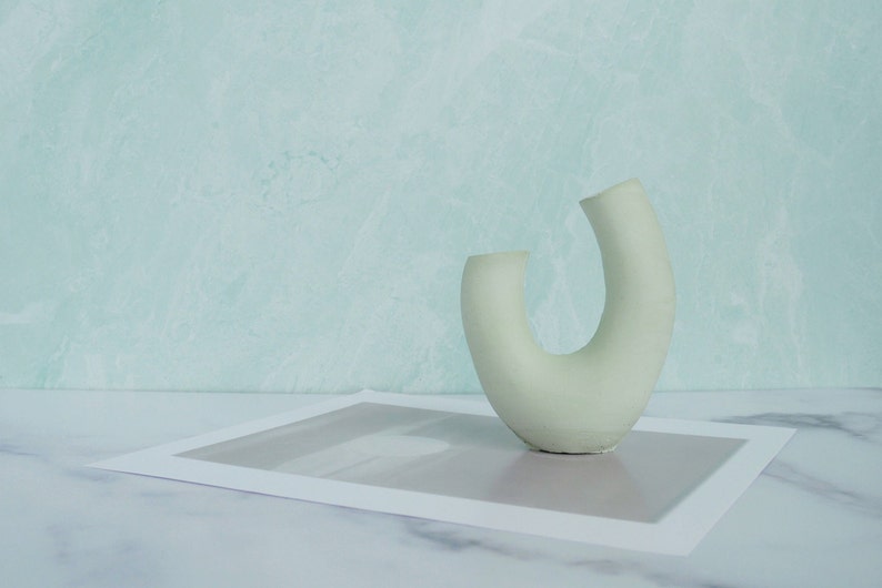 Minimalist product photography prop showcasing a single modern U-shaped plaster arch vase. The handmade concrete styling block features an elegant, asymmetrical curved ring silhouette in a soft, pale sage green. It stands on a white marble surface over a gray abstract art print card, set against a soft, textured pastel teal wall background. A sleek, contemporary display riser ideal for showcasing aesthetic items, jewelry, or home fragrance styling.
