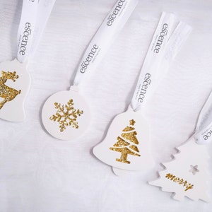 May include: Four white Christmas ornaments with gold glitter accents. The ornaments feature a reindeer, a snowflake, a Christmas tree, and a Christmas tree with the word "merry" on it. Each ornament has a white ribbon with the word "essence" printed on it.