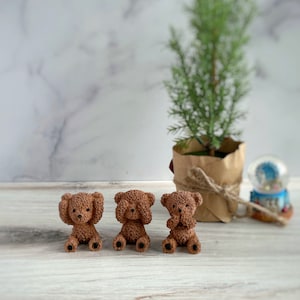 May include: Three brown teddy bear figurines, each with a different hand gesture: one covering its eyes, one covering its ears, and one covering its mouth. The figurines are arranged in a row on a light wood surface.