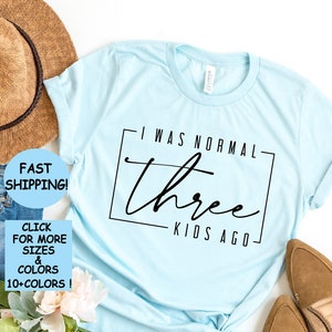 Puede incluir: Una camiseta azul claro con el texto "Yo era normal hace tres hijos" impreso en ella. La camiseta está colocada sobre una superficie blanca con un par de zapatos marrones, un sombrero de paja y un par de pantalones vaqueros azules.