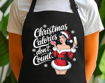 Puede incluir: Delantal negro con un bolsillo grande, con un gráfico temático navideño. El gráfico incluye una pin-up con un traje de Papá Noel, sosteniendo una rosquilla y champán, con el texto "Christmas Calories don't Count."
