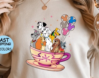 May include: A white sweatshirt with a graphic of a teacup filled with Disney's 101 Dalmatians characters. The teacup has a pink and white floral design. The characters are all smiling and looking up at the viewer. There are also several colorful balloons in the background.