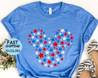 Può includere: Una maglietta blu con una silhouette di Topolino rosso, bianco e blu fatta di stelle. La maglietta è su uno sfondo bianco con un paio di scarpe a righe e un cerchio blu con il testo "Heather Columbia Blue".