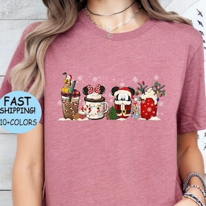 以下が含まれることがあります： ヘザーモーヴのTシャツに、クリスマスをテーマにしたミッキーマウス、ミニーマウス、グーフィーがホリデーをテーマにしたカップを持っているデザインが施されています。デザインには雪片とクリスマスライトが含まれています。