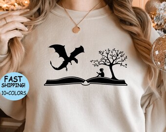 May include: A cream-colored sweatshirt with a black graphic of a dragon flying over an open book with a tree and a person sitting under the tree. The text "FAST SHIPPING! 10+ COLORS" is on a blue circle.