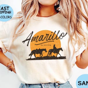 May include: Sand-colored t-shirt featuring the text "Amarillo by morning" above a silhouette of a horse and rider against an orange sun. The shirt also has the text "Fast Shipping! 10+ Colors" in a blue circle.