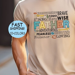 Op de afbeelding: Een beige T-shirt met een grafisch ontwerp met het woord "Vader" in een kleurrijke marquee-stijl. De grafiek bevat ook woorden als "hardwerkend", "moedig", "grappig", "wijs", "sterk", "avontuurlijk", "onverschrokken", "toegewijd", "beschermer" en "liefhebbend".