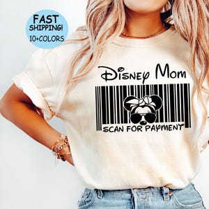 以下が含まれることがあります： 「Disney Mom」と「Scan for Payment」と書かれた白黒のバーコードデザインの白いTシャツ。バーコードの中に白黒のミッキーマウスの漫画の画像があります。シャツは青いジーンズを履いたモデルが着ています。