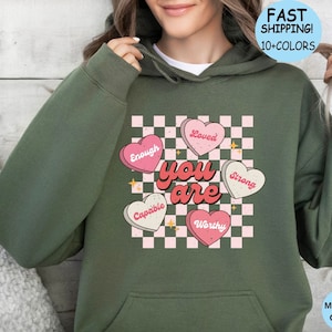 May include: Green hooded sweatshirt with a checkered pattern and pink hearts that say "you are" with words like "enough", "loved", "capable", "worthy", and "strong" inside the hearts.