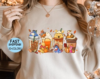 May include: A beige sweatshirt with a graphic of cartoon characters holding cups of coffee, tea, and hot chocolate. The characters are Winnie the Pooh, Tigger, Piglet, and Eeyore. The graphic is surrounded by fall-themed elements like leaves, pumpkins, and acorns.