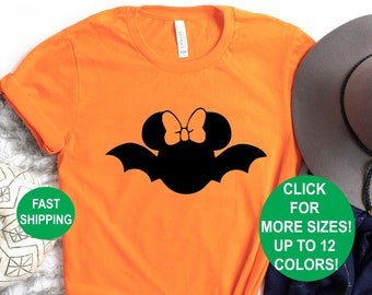 Puede incluir: Camiseta naranja con una silueta de murciélago negro con un lazo en la parte superior. El murciélago es una versión de un personaje de dibujos animados con temática de Halloween. La camiseta está rodeada de decoraciones de Halloween, incluyendo una telaraña, calabazas y un sombrero.