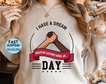 May include: A beige sweatshirt with the text "I Have A Dream" and "Martin Luther King Jr. Day" in a red, white, and black graphic design. The graphic features two hands clasped together.